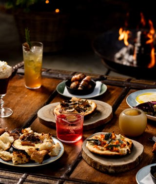 A portrait image a selection of fire-cooked dishes and cocktails from the Fire Garden menu at Ham Yard Hotel, they sit on a wooden coffee table