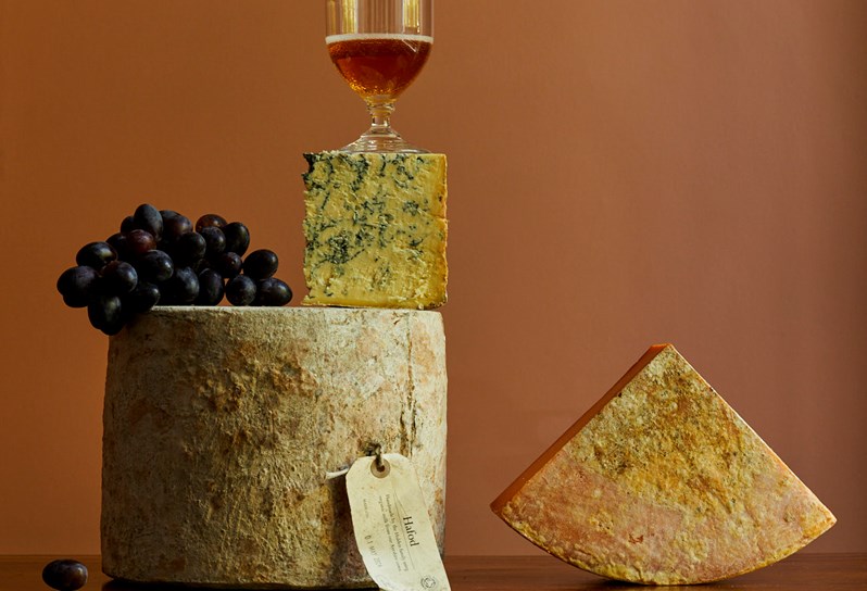 Collection of cheeses, with grapes perched on top of barrel shaped cheese, and wine balancing on top.