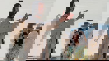 Still from the film 'Good Fortune'. A man dressed in a trench coat with angel wings stands with an arm outstretched looking off screen. Below him, a woman with angel wings looks up at him