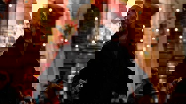 A still from "Shelter" showing a man wearing a black coat. Bright background of lights behind.