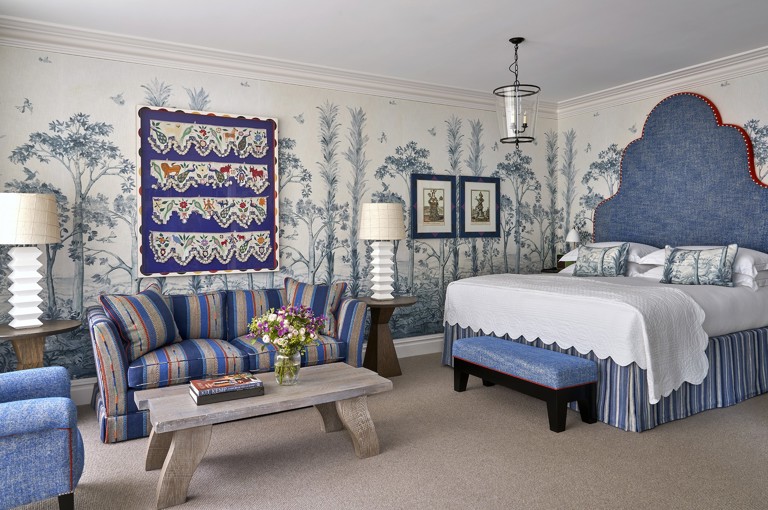 A diagonal shot of the bedroom in a Deluxe Junior Snug Suite at Ham Yard Hotel, featuring a king bed with a large headboard, a blue sofa, armchair and coffee table with two small side tables and lamps either side