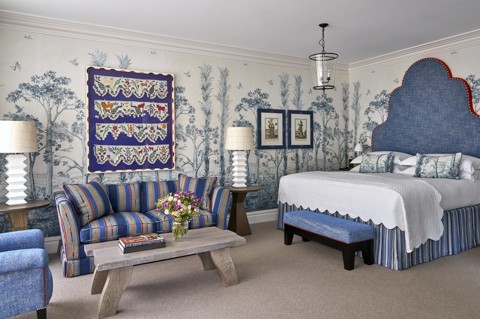 A diagonal shot of the bedroom in a Deluxe Junior Snug Suite at Ham Yard Hotel, featuring a king bed with a large headboard, a blue sofa, armchair and coffee table with two small side tables and lamps either side