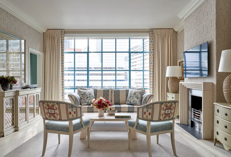 A living room area. Two geometric patterned and blue Whitby chairs in front of a coffee table opposite a blue and white striped sofa with floor to ceiling windows behind. To the left, a cream cabinet unit with a large frame handing above. To the right, a fireplace and TV with chest of drawers and lamps either side.