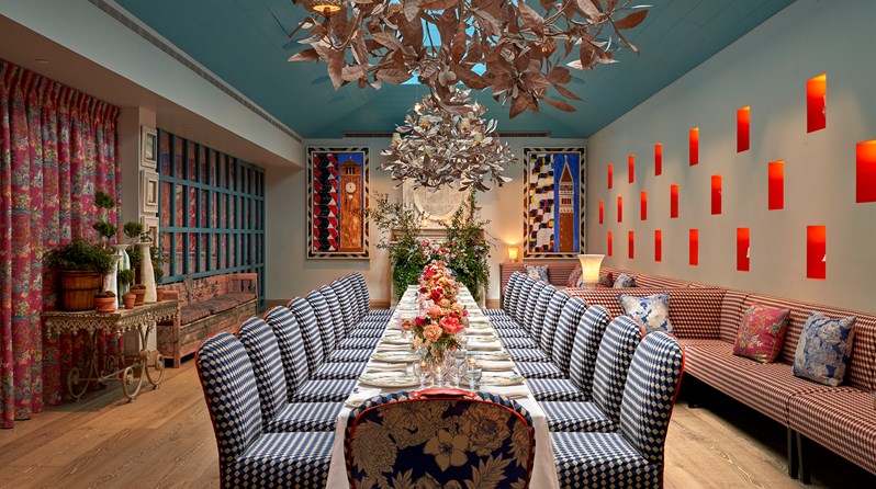 The Orangery at Warren Street Hotel with a vaulted blue ceiling and skylights. Banquette seating runs down the right-hand side of the room and a long table runs down the centre of the room with chairs upholstered in a blue, white and red patterned fabric. Two statement chandeliers hang from the ceiling and at the end of the room is a fireplace filled with foliage.
