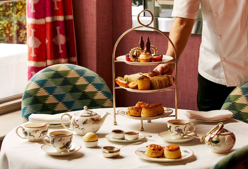 A three-tiered London Calling themed afternoon tea. The first tier contains desserts such as The Guards blackberry macaron, London Eye cinnamon sablé etc .You can see the hand of the chef placing it on the table. There are finger sandwiches as well as Chicken Scotch egg. The bottom tier contains four freshly baked scones with clotted cream and jam on the side. Accompanying on the table, there is a tea as well as two plated scones.