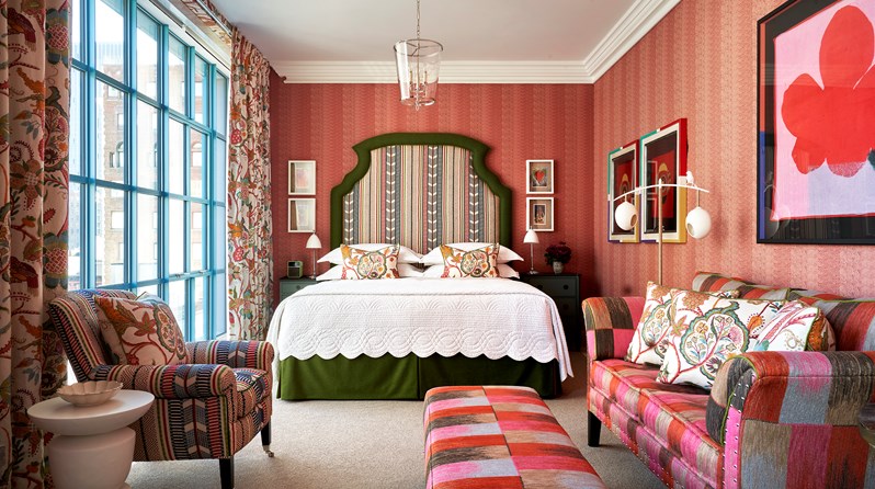 A dark green bed with a headboard upholstered in a muted striped fabric, a matching patterned arm chair on the right of the room with a white side table next to it. A Willow Ikat weave sofa and footstall on the left.