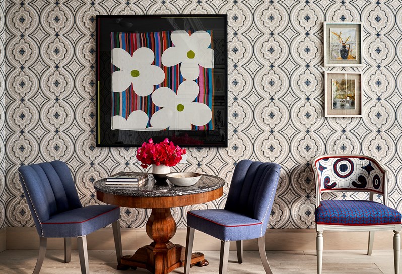 Two dark blue chairs either side of a marble table with pink flowers on, with a multi-coloured and white flower print hanging on the wall above. On the right, a blue and red patterned chair below two small paintings hanging on the wall.