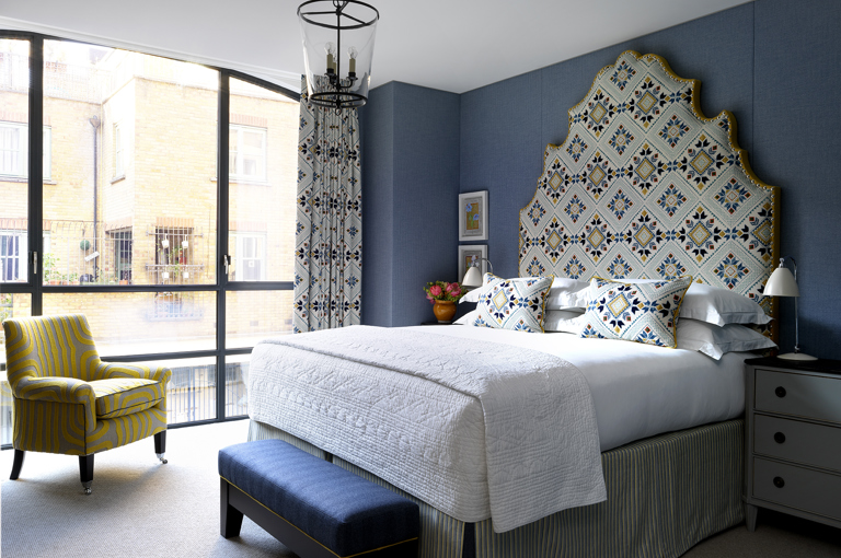 A wide shot of the view of the one bedrooms , A large statement headboard and bed dressed in crisp white sheets rest against walls upholstered in a dark navy fabric. There are floor to ceiling windows and a patterned yellow armchair in front of the windows at 9-11 Richmond Buildings.