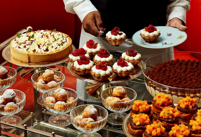 An image to promote the Long Lunch at Ham Yard Bar & restaurant at Ham Yard Hotel, The image shows a luxurious dessert trolley laden with an assortment of intricately presented pastries and cakes. A server in formal attire is plating a raspberry tart.