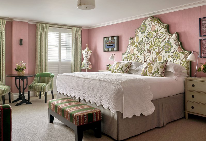 Diagonal bedroom shot a deluxe room, 005, at Knightsbridge Hotel with plain light pink walls and green nature drawings on the headboard, pillows and mannequin. There is also two chairs and a desk as well as a sofa and a long chair.