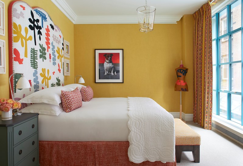 A yellow bedroom with a large multicoloured headboard, grey bedside table and yellow footstool
