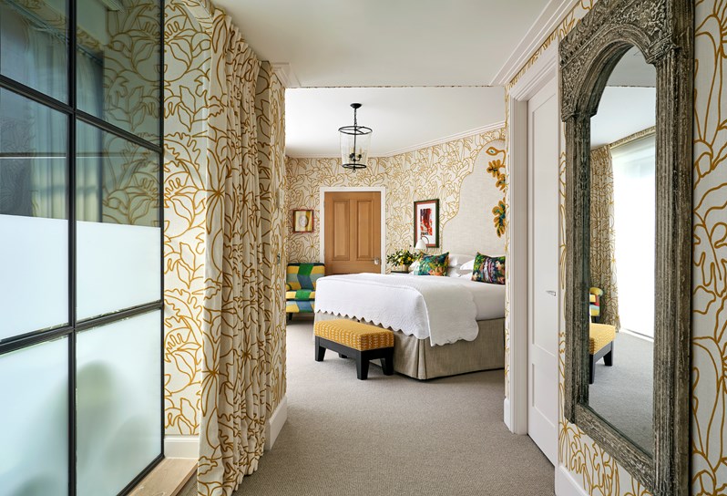 A shot looking down a corridor into a bedroom with walls upholstered in a lining embroidered with a yellow floral outline. A double bed has a large headboard with a leaf motif embroidered onto it. There is a yellow footstall at the end of the bed and a multicoloured 'Zig Zag' chair to the left.