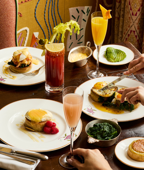 A portrait shot of brunch dishes being served in the Dora Carrington Room at Charlotte Street Hotel: there are various dishes using crumpets as well as cocktails on the table