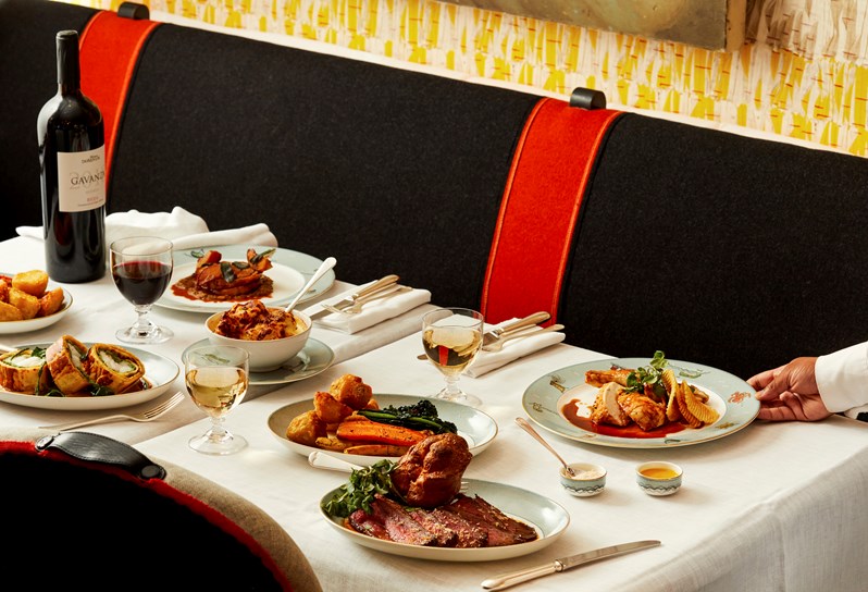 The Long Lunch at Ham Yard Bar & Restaurant at Ham Yard Hotel. The image shows a table set with an array of dishes, including roast beef with Yorkshire pudding, seasonal vegetables, and potatoes. Glasses of red and white wine and a bottle of red wine are on the table. A server is placing a plate of roasted chicken with crisp potatoes.