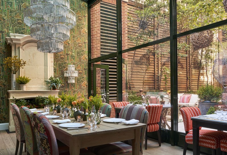 Diagonal angle shot of the Orangery facing the corner with the curtain wall on the right and the fire place and chandeliers wall on the back left. There three tables set up including one large table in the middle of the room and front left of the shot.