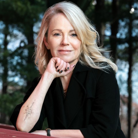 A headshot of author and podcaster Dani Shapiro. She is stood outside, resting her chin on her arm leaning on a wooden beam. Trees can be seen out of focus behind her