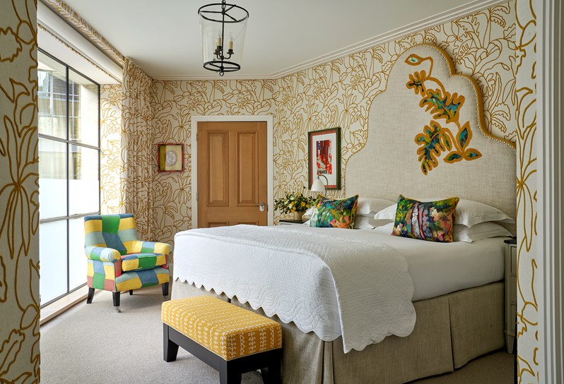 A bedroom with walls upholstered in a lining embroidered with a yellow floral outline. A double bed has a large headboard with a leaf motif embroidered onto it. There is a yellow footstall at the end of the bed and a multicoloured 'Zig Zag' chair to the left.