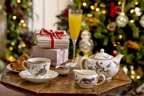 This image shows a festive breakfast setting at Haymarket Hotel, featuring mythical creature crockery with tea, croissants, preserves and a glass of orange juice, set beside colourful wrapped presents and a twinkling Christmas tree.