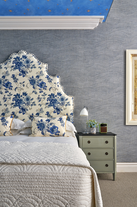A portrait image focusing on the bed in The Knightsbridge Suite at Knightsbridge Hotel. In this image we see the bed with white sheets and pillows and decorative white and blue statement headboard with matching decorative pillows. To the right, we see a green bedside table with shelves.