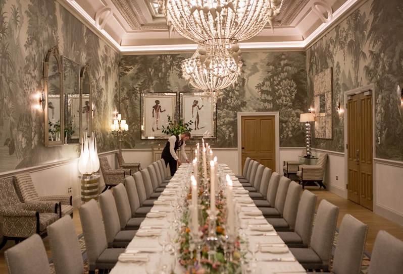 A wide shot looking down a long table dining set up in the Shooting Gallery at Haymarket Hotel. The table is dressed with colourful bouquets of flowers and long candle sticks. A waiter lays a place setting at the far end of the table