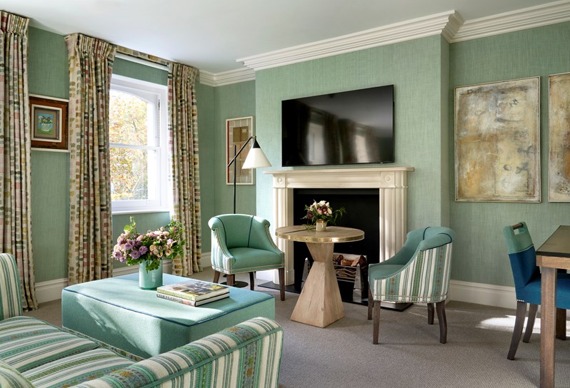 Diagonal bedroom shot the lounge in a junior suite with plain light blue walls and of a sofa, two chairs and a long chair in blue, beige and green stripes. There is also a fire place and a television