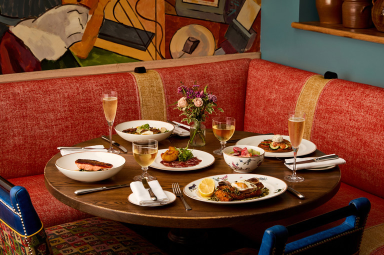An image to promote Oscar Restaurant’s À La Carte menu at Charlotte Street Hotel. The image shows a round dark wood table with multiple dishes, examples of dishes include chicken schnitzel and trout dishes and with flutes of champagne and glasses of wine on the table.
