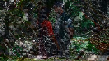 A still from 'Hamnet'. A man and woman dressed in tattered period clothing stand close face to face with one another in the middle of the woods