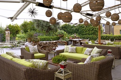 Diagonal shot from the bar of the Roof Terrace at Ham Yard Hotel, it shows the lime green cushioned and wicker sofa seating area with a coffee table in the centre. 