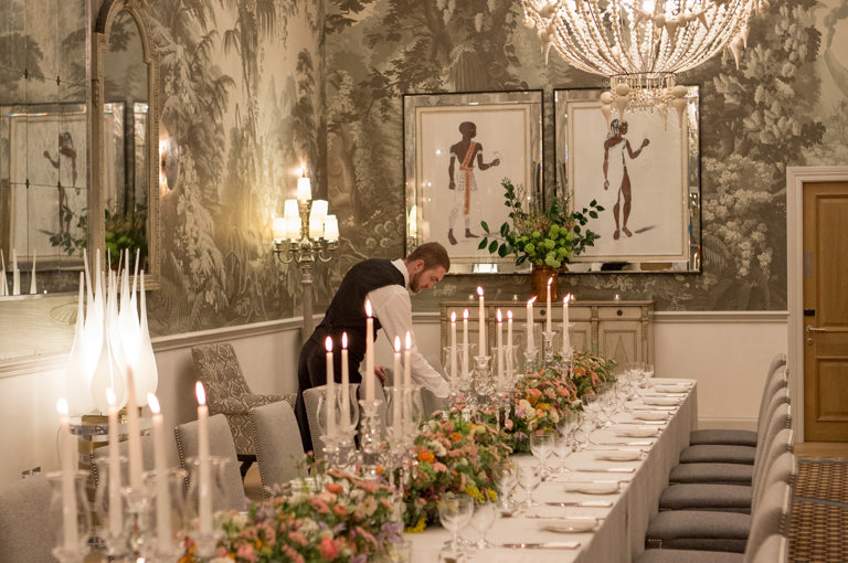 Portrait shot of a long dining table dressed with colourful flowers and tall candlesticks with a waiter at the end, in the Shooting Gallery private events room at Haymarket Hotel
