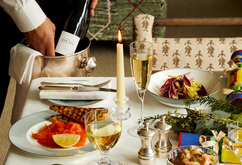 A portrait closeup of a table setup for festive dining. There are Christmas crackers, candles, and plates with starters being laid out, alongside a glass of white wine and champagne. The starters include, smoked salmon, autumn leaves salad, amongst others. The crockery being used is Mythical creatures.