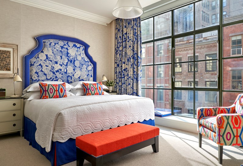 Diagonal shot of the bedroom in a Deluxe Room in The Crosby Street Hotel with a large window on the right and a large bed with blue with flower patterns oversized headboard and matching curtains. Orange long chairs at the bottom of the bed. An arm chair on the right with matching colourful patterns to the bed cushion