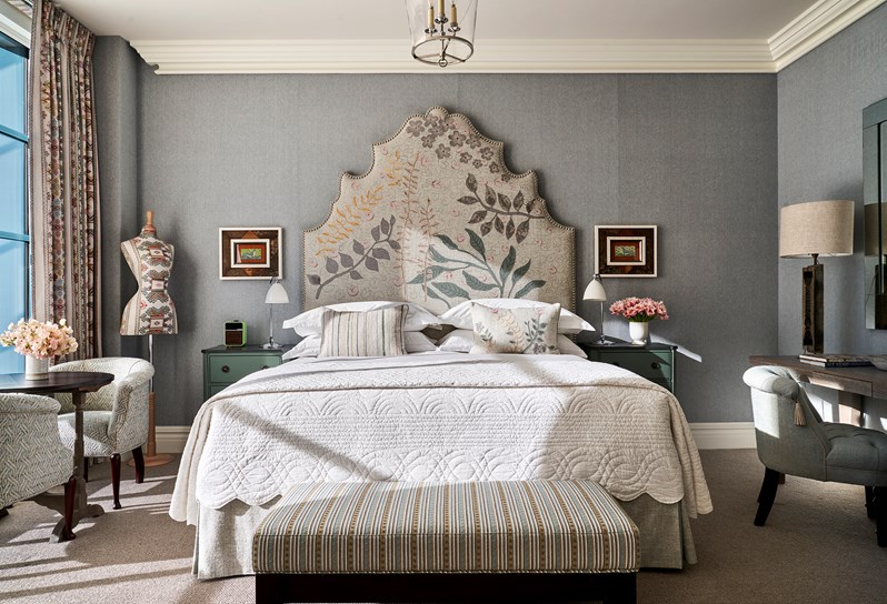 A bed with a neutral coloured floral pattern. To the left, geometric patterned mannequin, two blue patterned armchairs and a coffee table with pink flowers on. To the right, a blue cushioned chair in front of a desk with a beige lamp on