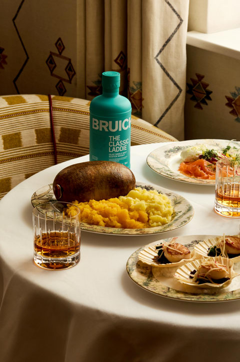 Portrait shot of a table set for Burns Night dinner with Bruichladdich at Covent Garden Hotel. There is a plate of haggis being cut open, plates of scallops and smoked salmon and glasses of whisky and Bruichladdich bottles.