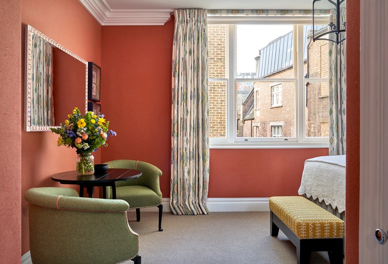 A view of the bed, the walls are a striking red colour and there is a green armchair witha black table in the middle with a beautiful flower bouquet on it and a yellow ottoman at the bottom of the bed.