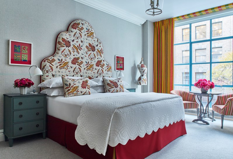Bed with red and orange patterned headboard and mannequin, orange curtains and two striped armchairs with a coffee table.