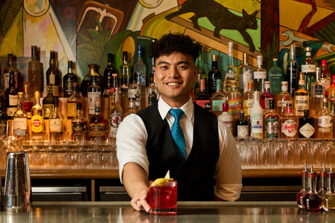 An image of a male bartender making the Ruby Reverie cocktail in the Refuel Bar, at Soho Hotel. The image shows the bartender smiling holding the finished cocktail.