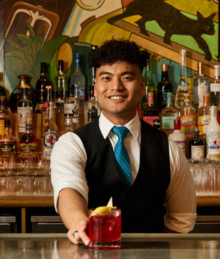 An image of a male bartender making the Ruby Reverie cocktail in the Refuel Bar, at Soho Hotel. The image shows the bartender smiling holding the finished cocktail.