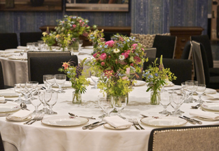 An close up image of the round dining table set up in the Indigo Room, there are white table cloths, floral bouquets in the centre and candles. each place around the table has a placemat, plate, cutlery, glassware and napkins ready for dining. private events room at The Soho Hotel.