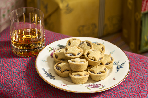 This image shows a festive Christmas setting at Haymarket Hotel, featuring a plate of traditional mince pies dusted with icing sugar, served alongside a glass of whisky, with colourful wrapped Christmas presents in the background.