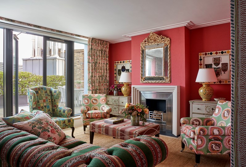 A living room space with coral red fabric walls and green and red patterned sofas and chairs. Art works hang either side of the fireplace and bi-fold doors open onto a terrace