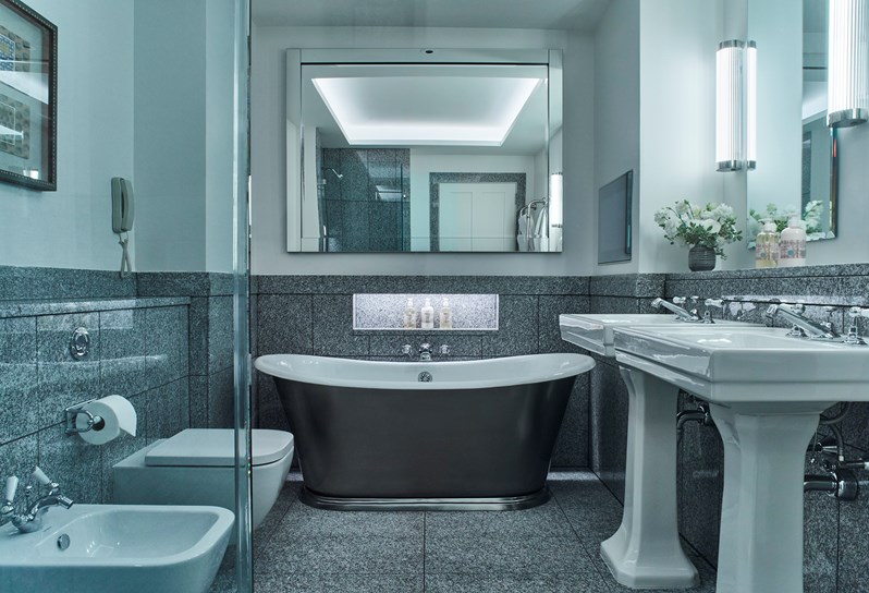 A grey marble bathroom, with a large metal bathtub, two parallel sinks in front of mirrors, a toilet and bidet