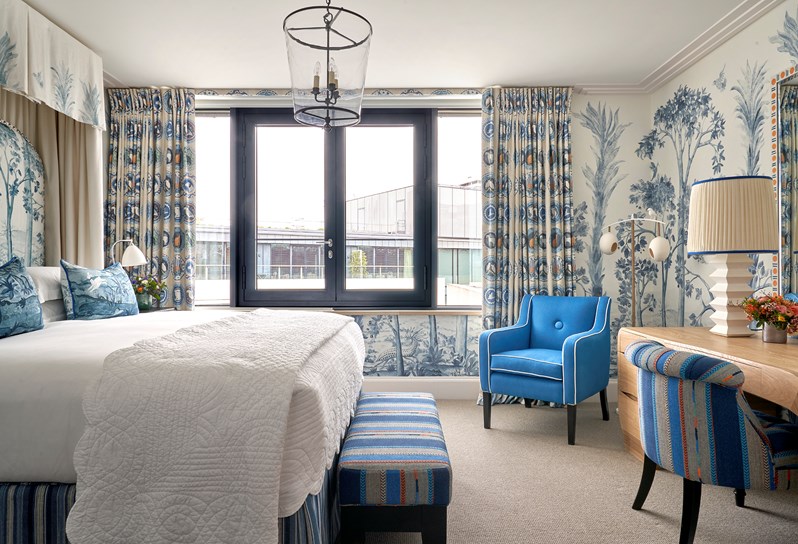 A canopied bed with a blue patterned headboard with blue cushions and a white valance. A blue and orange patterned footstool sits at the end of the bed and a wooden desk sits opposite the bed