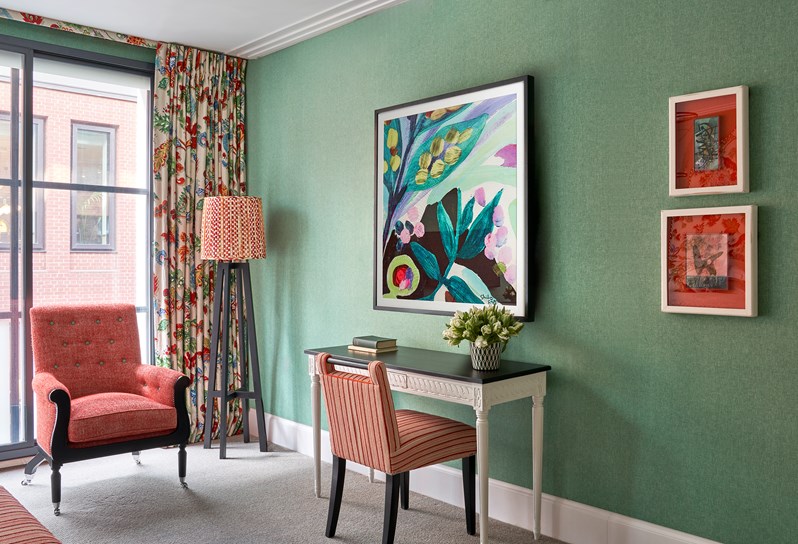 A seating area and original artwork in the bedroom with green wallpaper, as well as a with designed lamps, a chair and a  desk, and an artistic painting.
