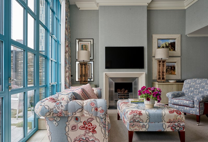 A living room area. A blue and white patterned armchair in front of footstall in blue 'Chainstitch' fabric which matches the sofa opposite and curtains on the floor to ceiling windows behind. There is a TV on the wall above the fireplace with two lamps on cabinets on either side.