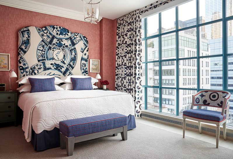 A bed with a blue and white patterned headboard, which matches the curtains and a Whitby chair by the windows. Blue and red cushions on the bed match the ottoman at the end.