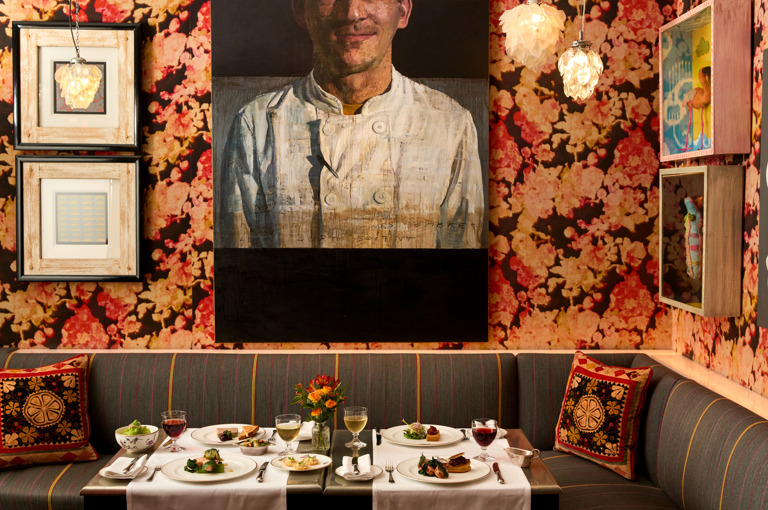 A image of a table at Refuel at Soho Hotel with white linen clothes is set up with multiple dishes from the Summer A La Carte with glasses of red and white wine on each place setting.