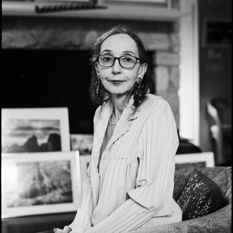 A black and white headshot of author Joyce Carol Oates. She is sat on a sofa in a lounge, artwork can be seen out of focus behind her
