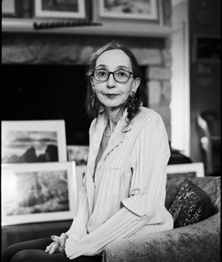 A black and white headshot of author Joyce Carol Oates. She is sat on a sofa in a lounge, artwork can be seen out of focus behind her