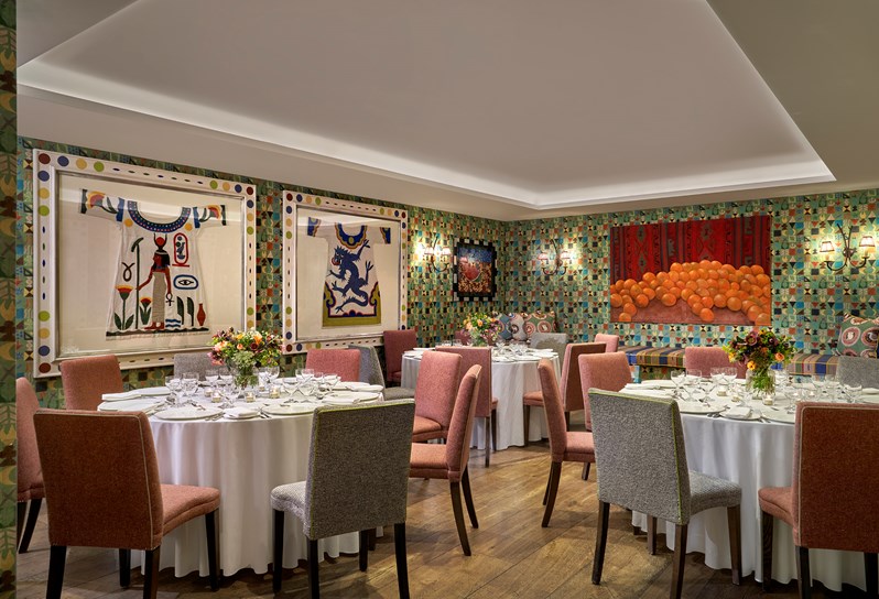 The newly photography Vanessa Bell Room setup for a dinner on round tables with orange chairs. There is beautiful bright artwork on the walls and flowers decorating each table.