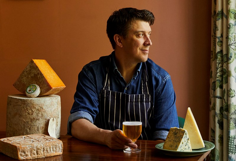 Portrait of Joe Fox sitting with cheeses on table, gazing into the distance